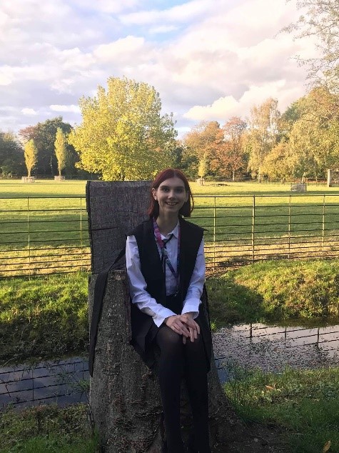 AJ (a red-haired girl) wearing subfusc and sitting on a tree-trunk chair after matriculation.