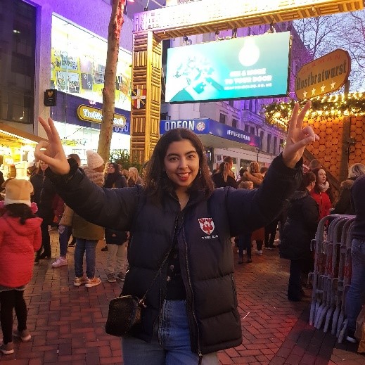 Aqsa standing with her hands up in peace signs and her tongue out. She wears a Queen's College puffer jacket and a crossover bag. She is standing in Birmingham Christmas market. You can see lots of people and stalls behind her, everything is lit up with Christmas lights.