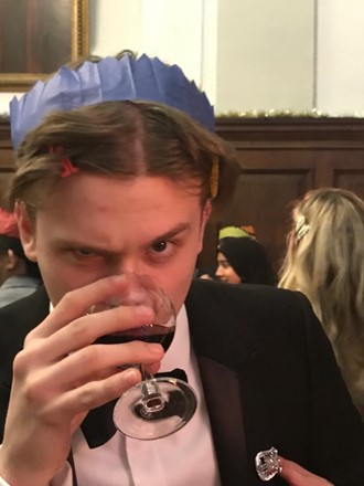 Luke at the Queen's Christmas Dinner, wearing a blue paper hat from a cracker and drinking a glass of red wine, while looking suspiciously at the camera.