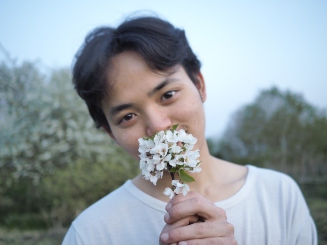 Mukahang in a white shirt, holding a bunch of white flowers which are covering his mouth.