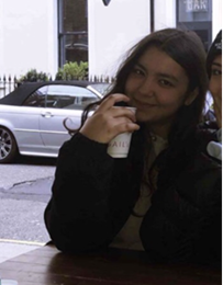 Sophie is smiling at the camera. She is in the right side of the frame and the background is bright behind her. You can see the street with some cars and a white building. She is drinking from a cup.