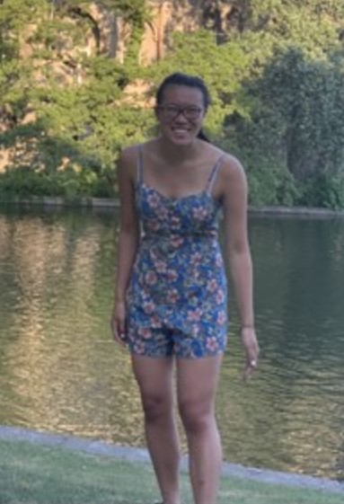 Ying in a blue playsuit with pink flowers standing on a river bank. She is smiling at the camera.